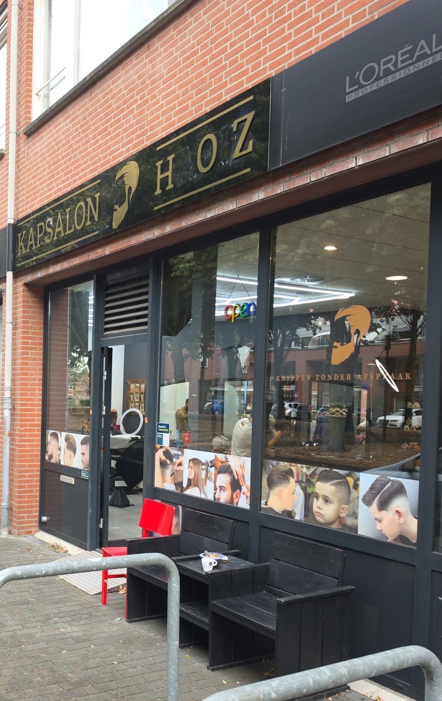 De gevel van Kapsalon HOZ met de zwarte naambalk en gouden letters, en de bankjes voor de deur